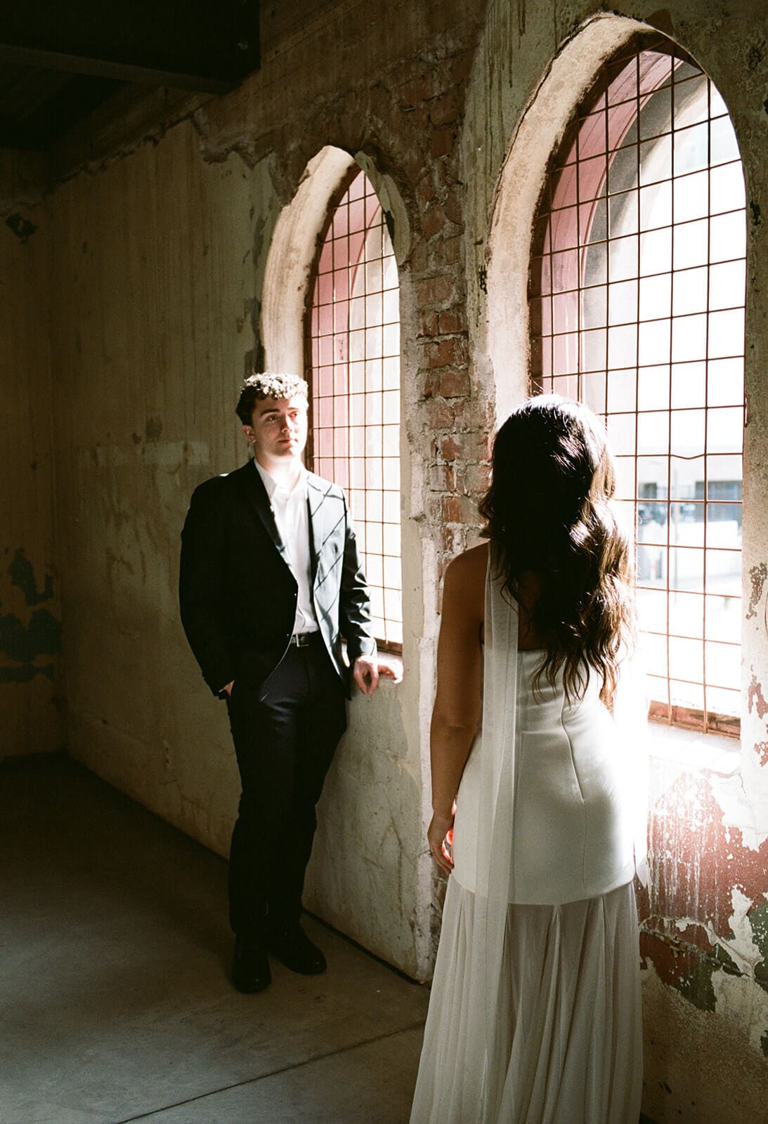 Bride and groom facing each other beside tall arched windows inside a historic venue.
