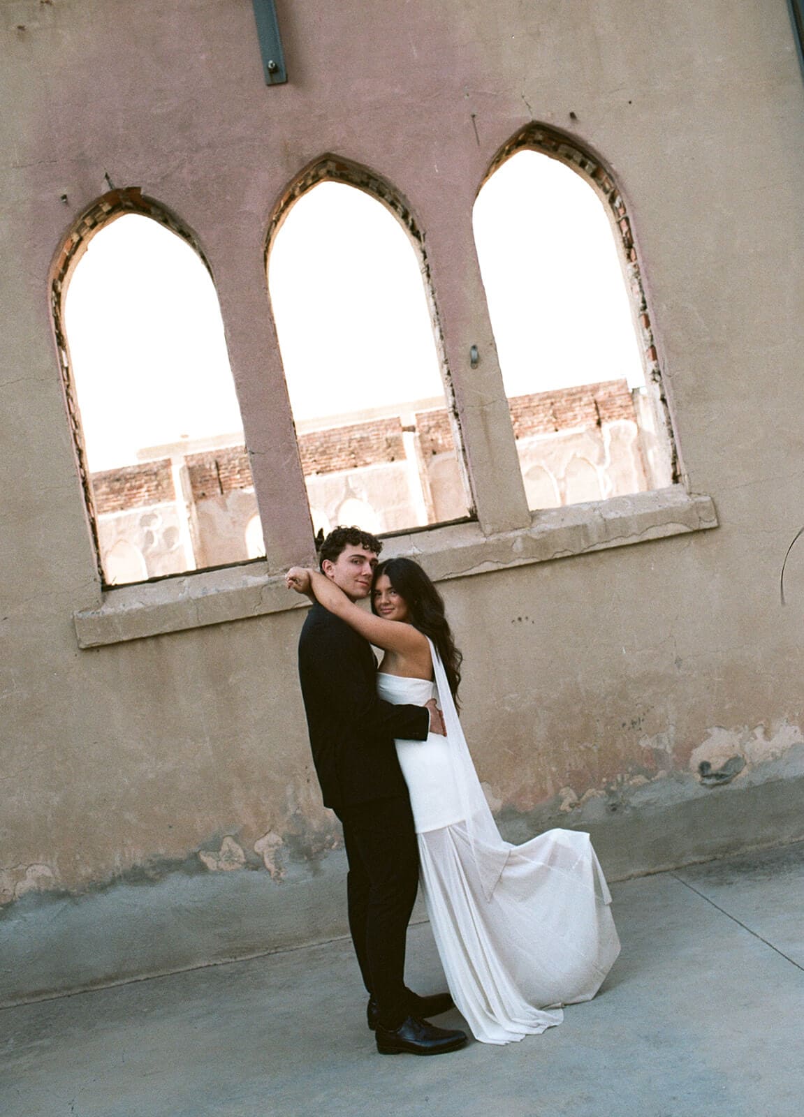 Newlyweds kissing outside a weathered building with arched windows.