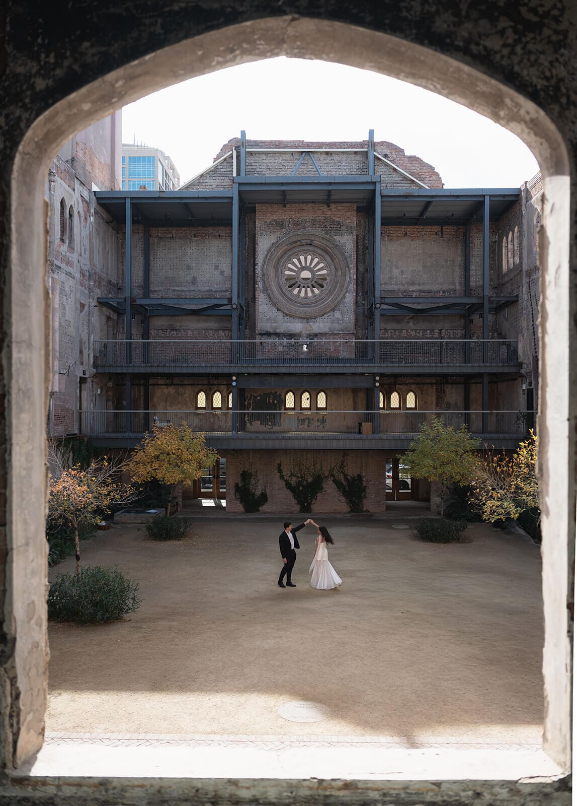 Couple standing in a courtyard framed by an arch and an ornate industrial facade.