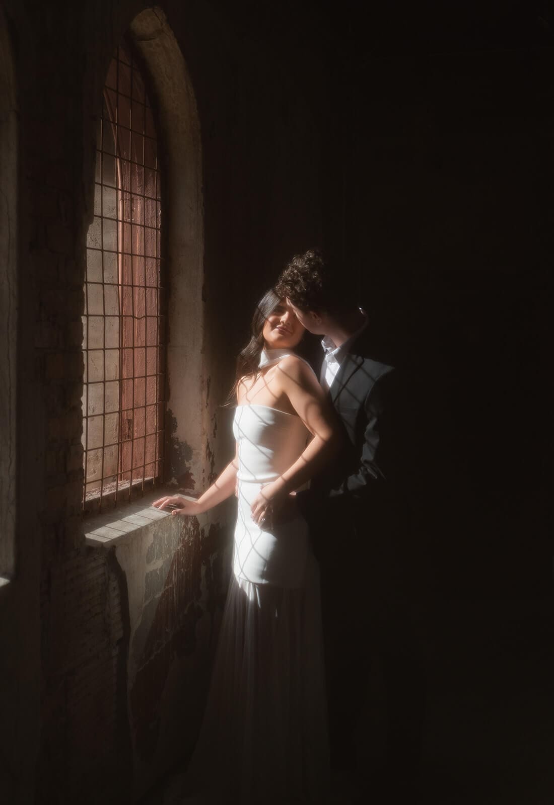 Bride and groom posing in a beam of sunlight beside a tall arched window.