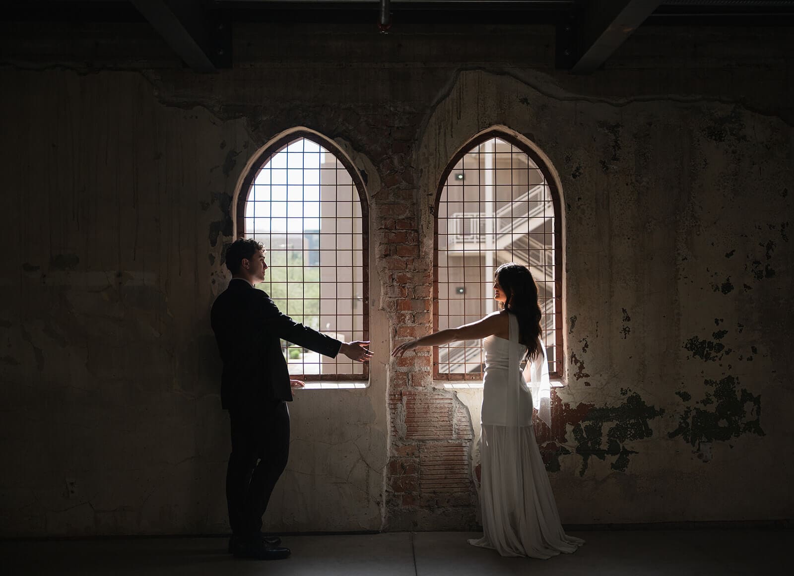 Bride and groom reaching toward each other in front of glowing arched windows inside an industrial wedding venue.