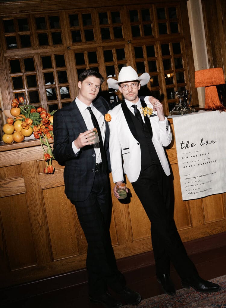 Two newlyweds in formal suits and cowboy hats holding drinks together inside a rustic reception space.