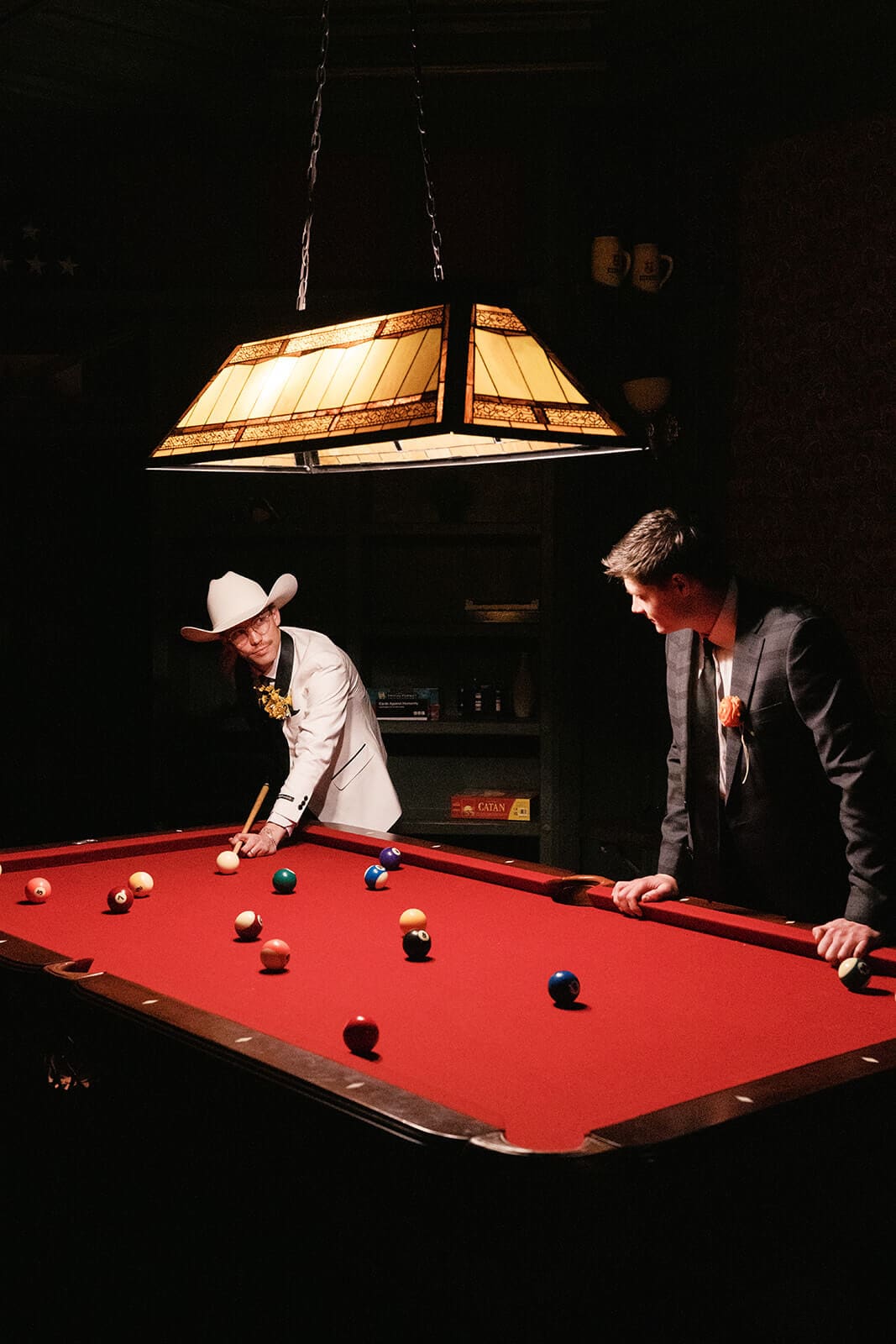 Two grooms playing pool beneath a hanging light inside a dark billiards room.