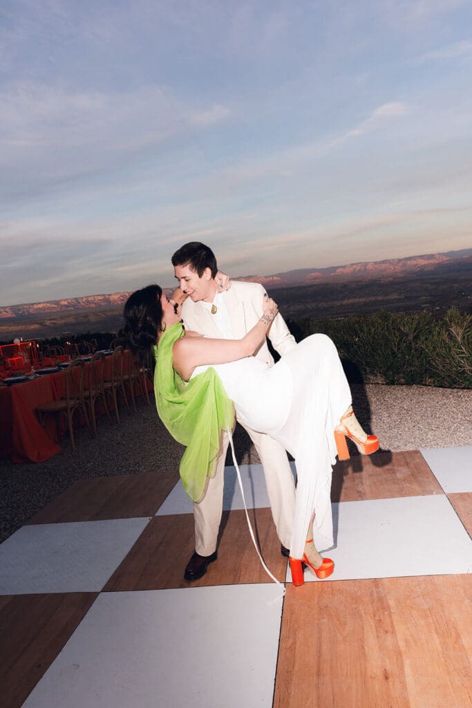 One newlywed carrying their partner across a checkered dance floor at sunset with mountains in the distance.