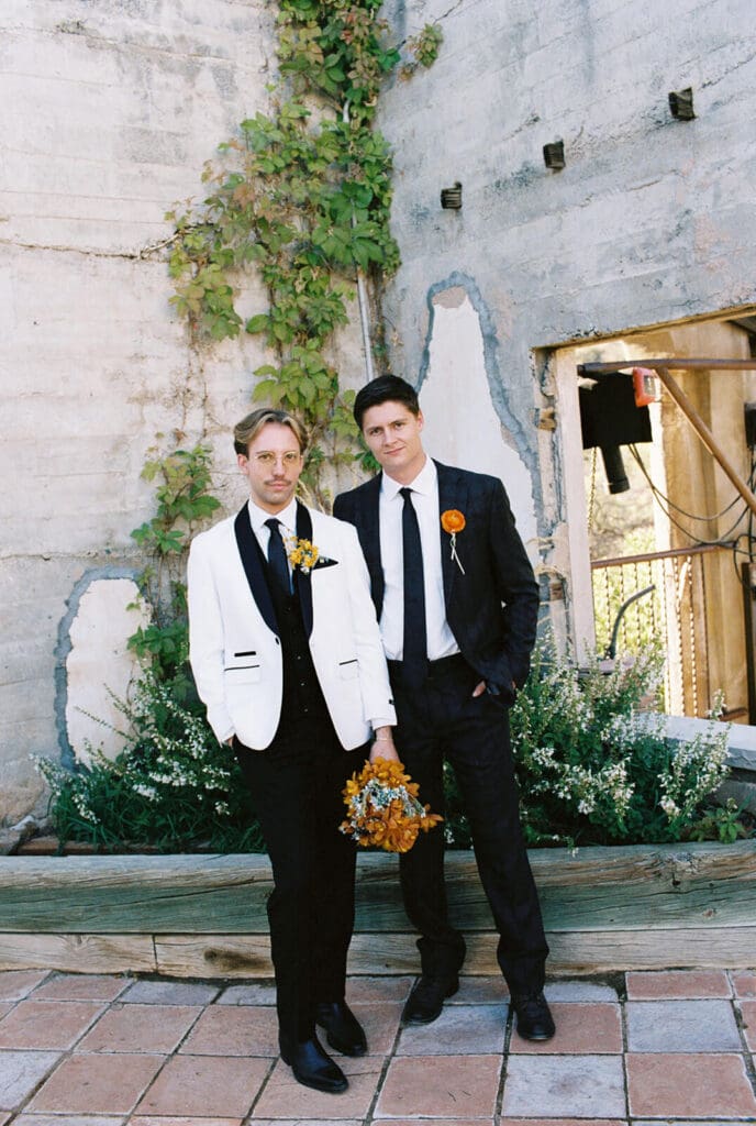 Couple in contrasting formal suits posing together against a weathered stone courtyard wall.