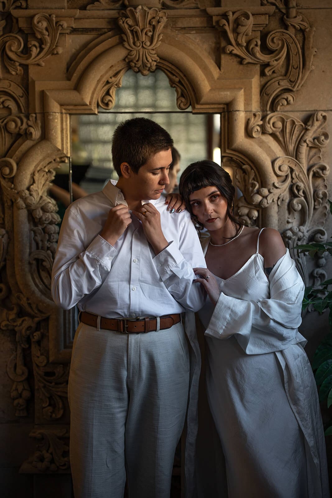 Partner buttoning their shirt collar as the bride stands close beside him in front of the same carved stone mirror, both dressed in white
