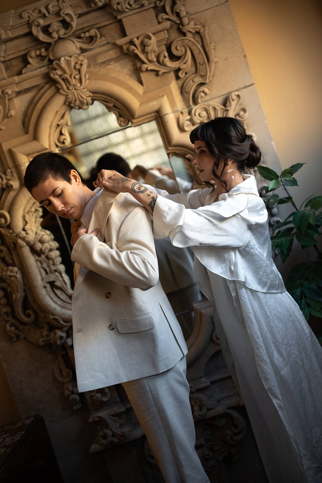 Bride adjusting her partner's suit jacket collar in front of an ornately carved stone mirror
