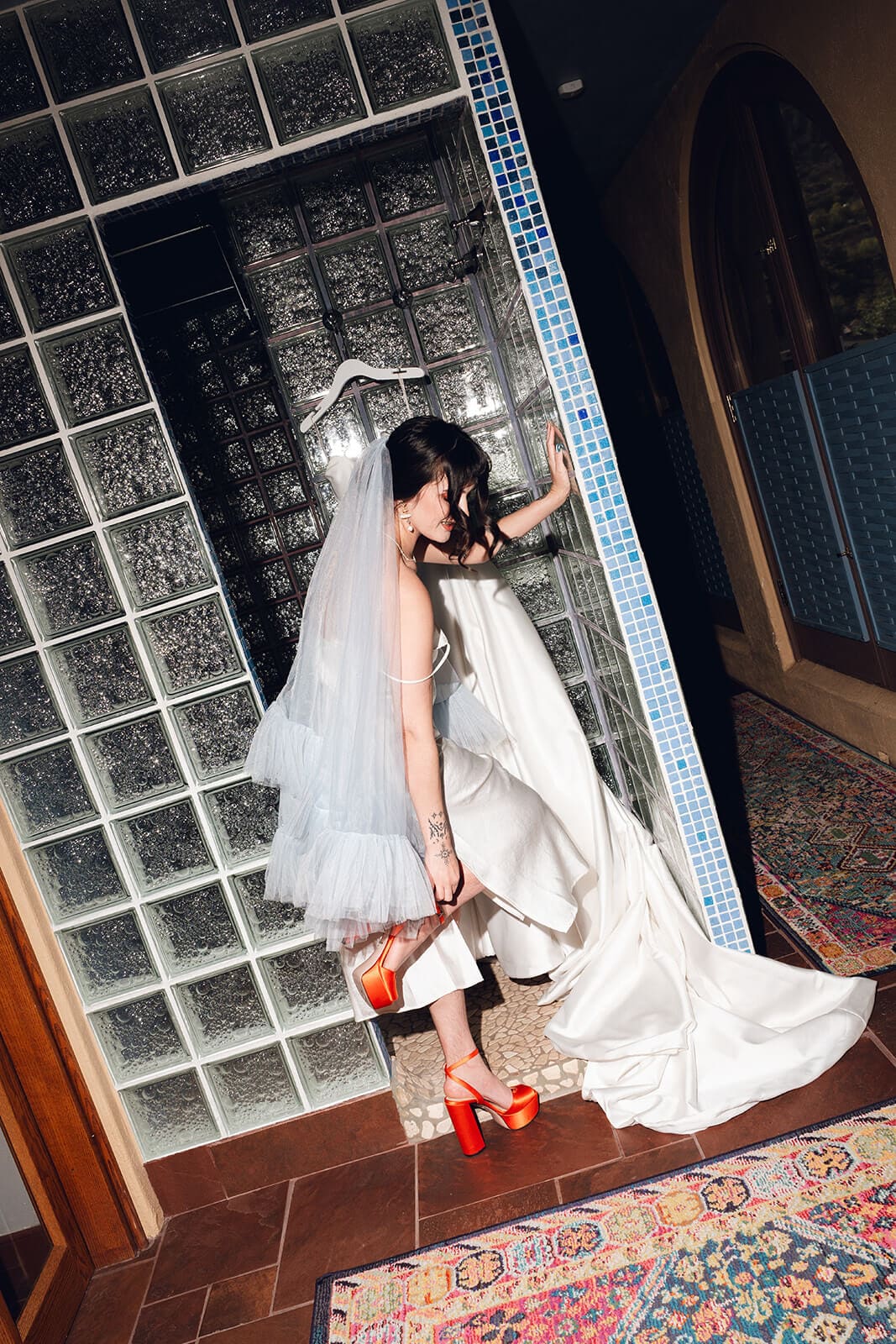 Bride in a white slip gown and tulle veil posing in the glass block doorway, balancing on one red platform heel
