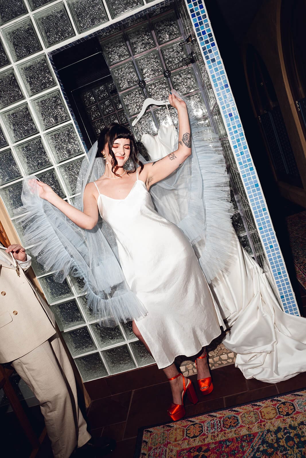 Bride dancing alone in a white slip gown and pale blue tulle veil that flares mid-motion, set against a glass block wall and colorful woven rug
