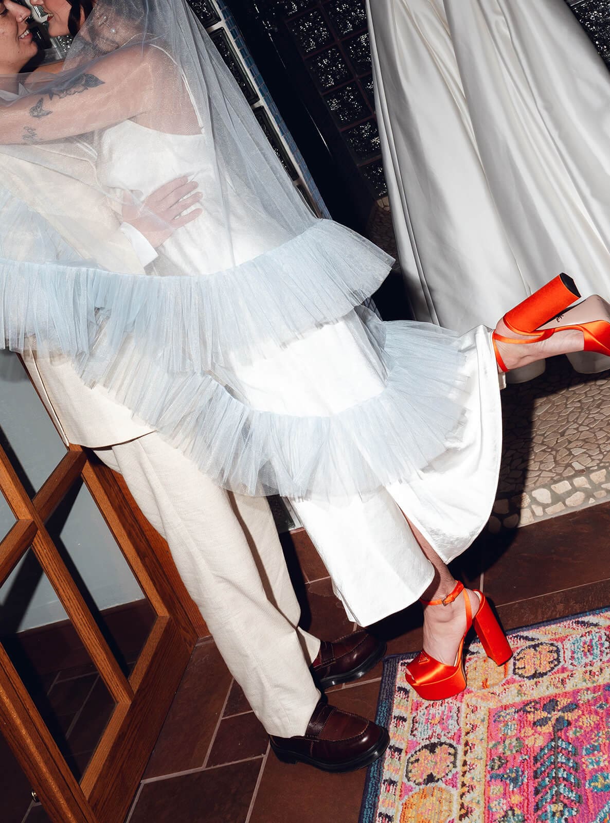 Close-up of the bride's tulle skirt layered over her partner's leg, her red platform heels crossed at the ankle

