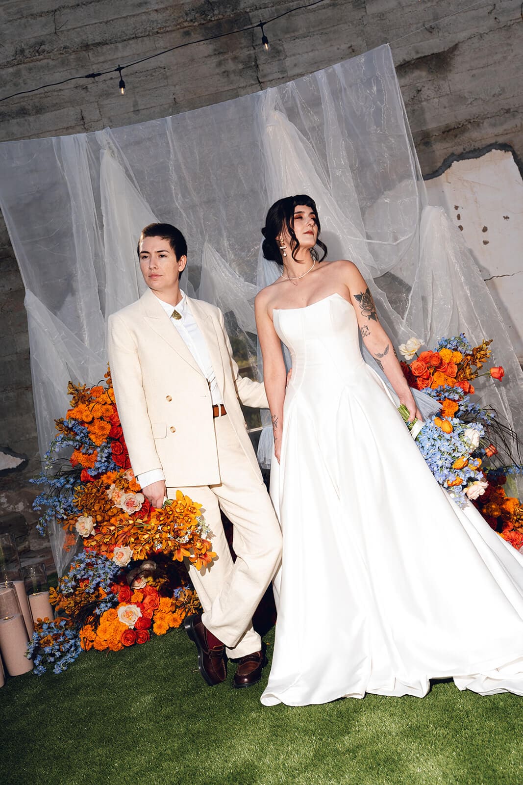 Couple standing in front of a large arrangement of orange and blue flowers with sheer draping cascading behind them
