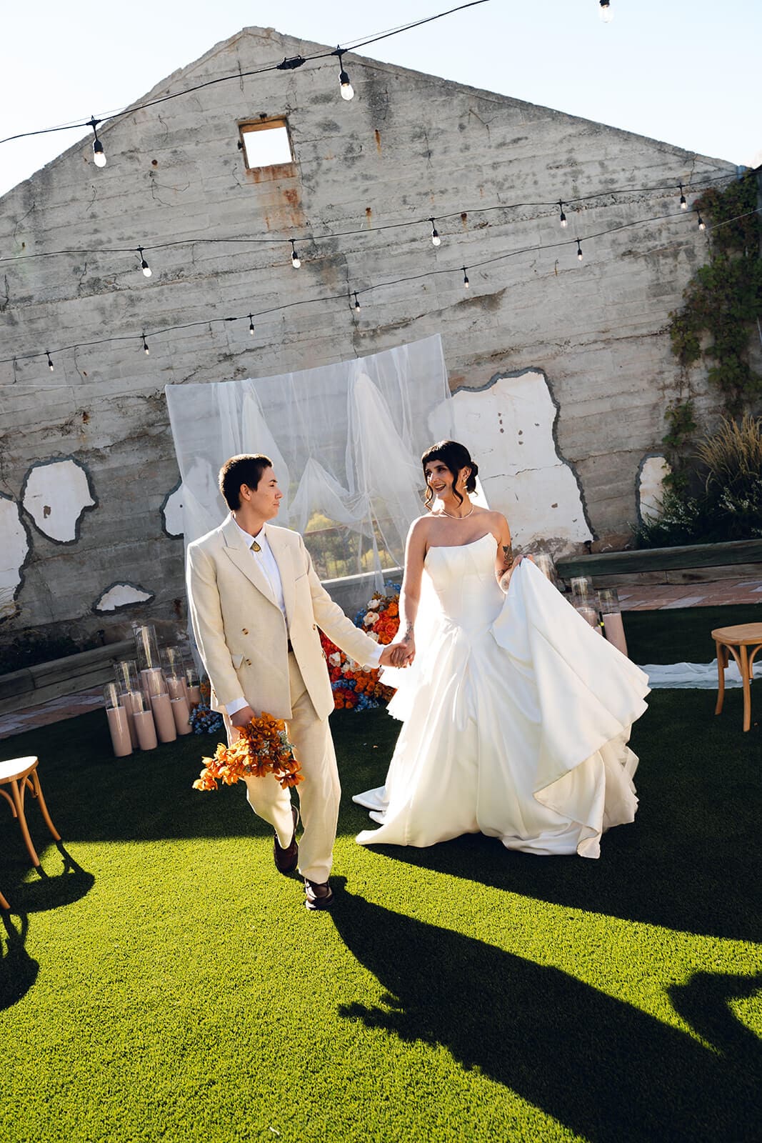 Couple walking hand in hand across the lawn at The Little Daisy, string lights strung overhead and a weathered concrete wall with painted patches behind them
