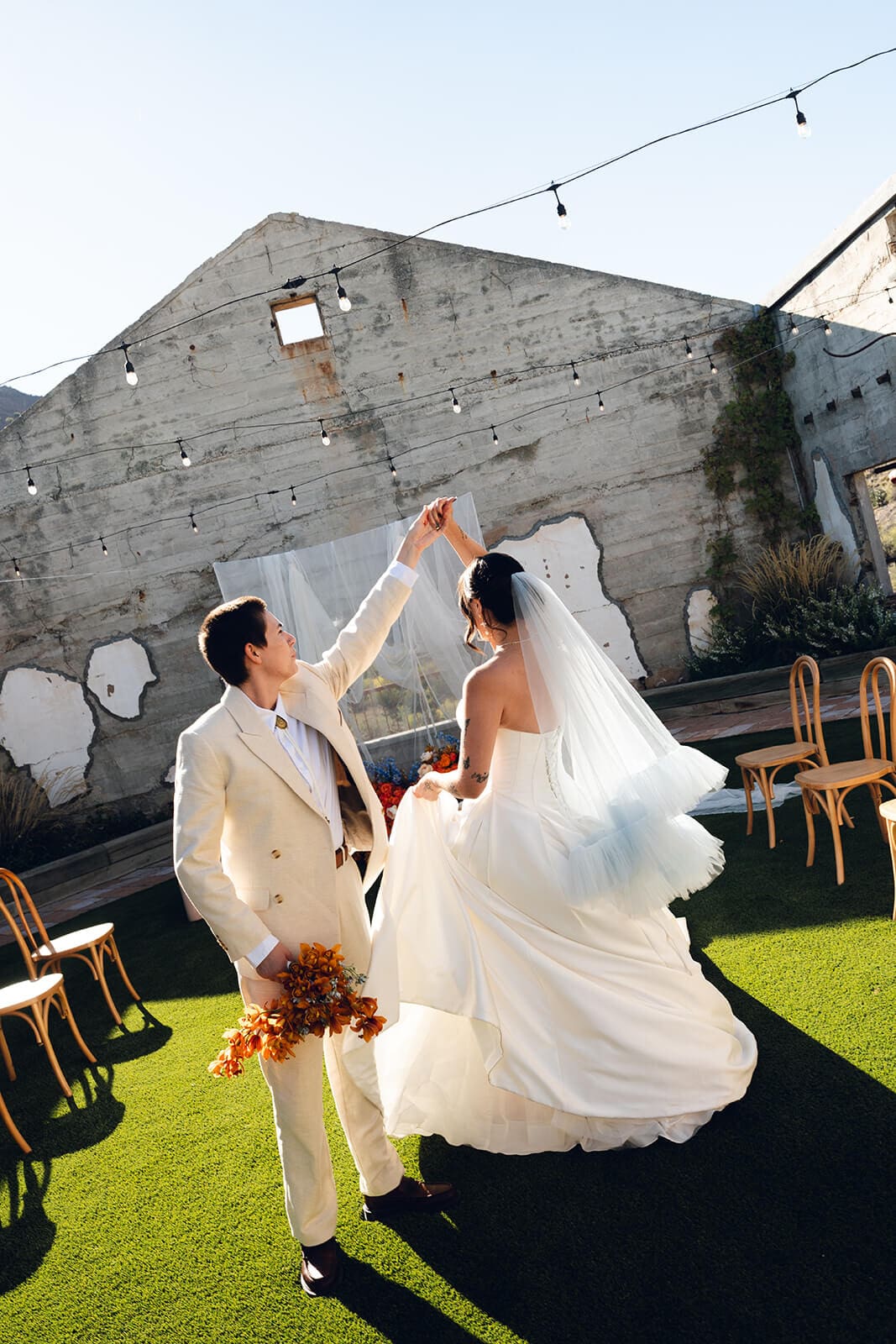 Couple embracing under string lights against a weathered concrete wall with peeling white patches, her veil catching the breeze and an orange bouquet in hand, wooden folding chairs lining the sunny lawn
