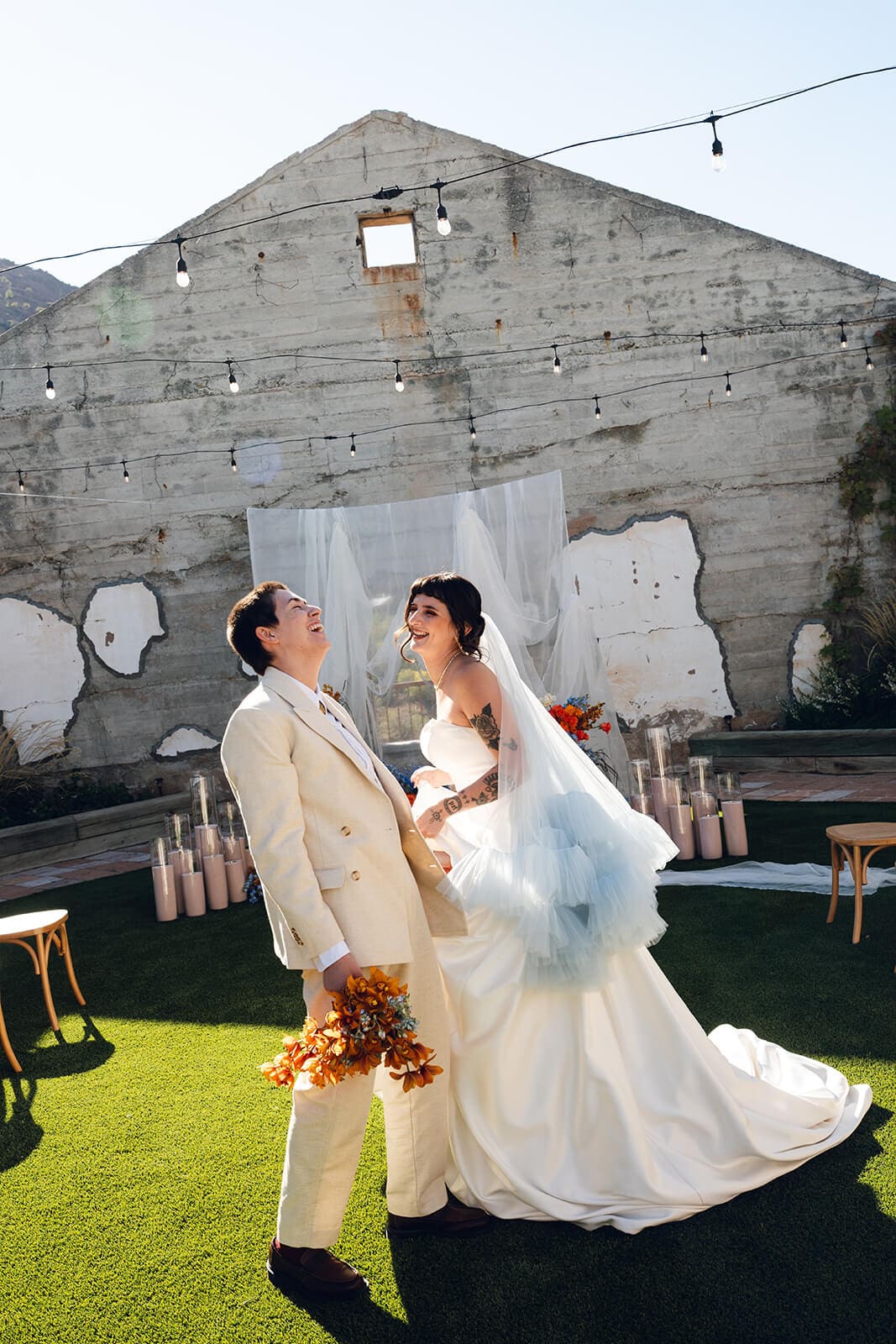 A couple walking hand in hand across the lawn at The Little Daisy, string lights strung overhead and a weathered concrete wall with painted patches behind them
