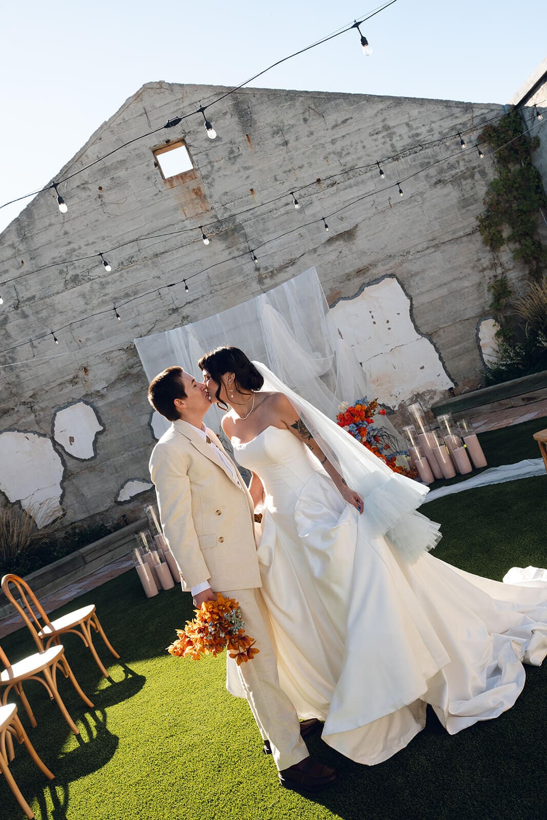 A couple lean in for a kiss, her pale blue veil swept sideways by the breeze, orange bouquet visible at her side

