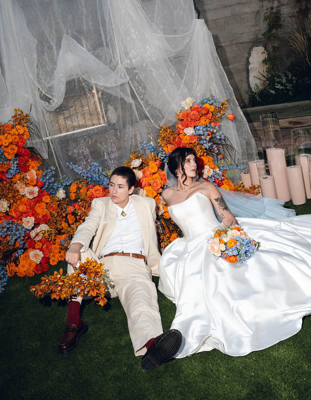 Bride and partner seated together on the grass, surrounded by a lush arrangement of orange and blue flowers with sheer draping overhead