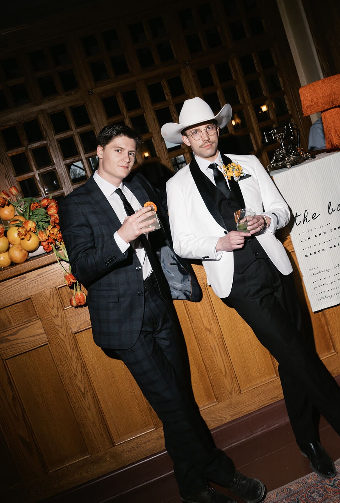 Two groomsmen holding drinks near a bar decorated with citrus fruit and orange floral arrangements, one wearing a white cowboy hat and yellow boutonniere
