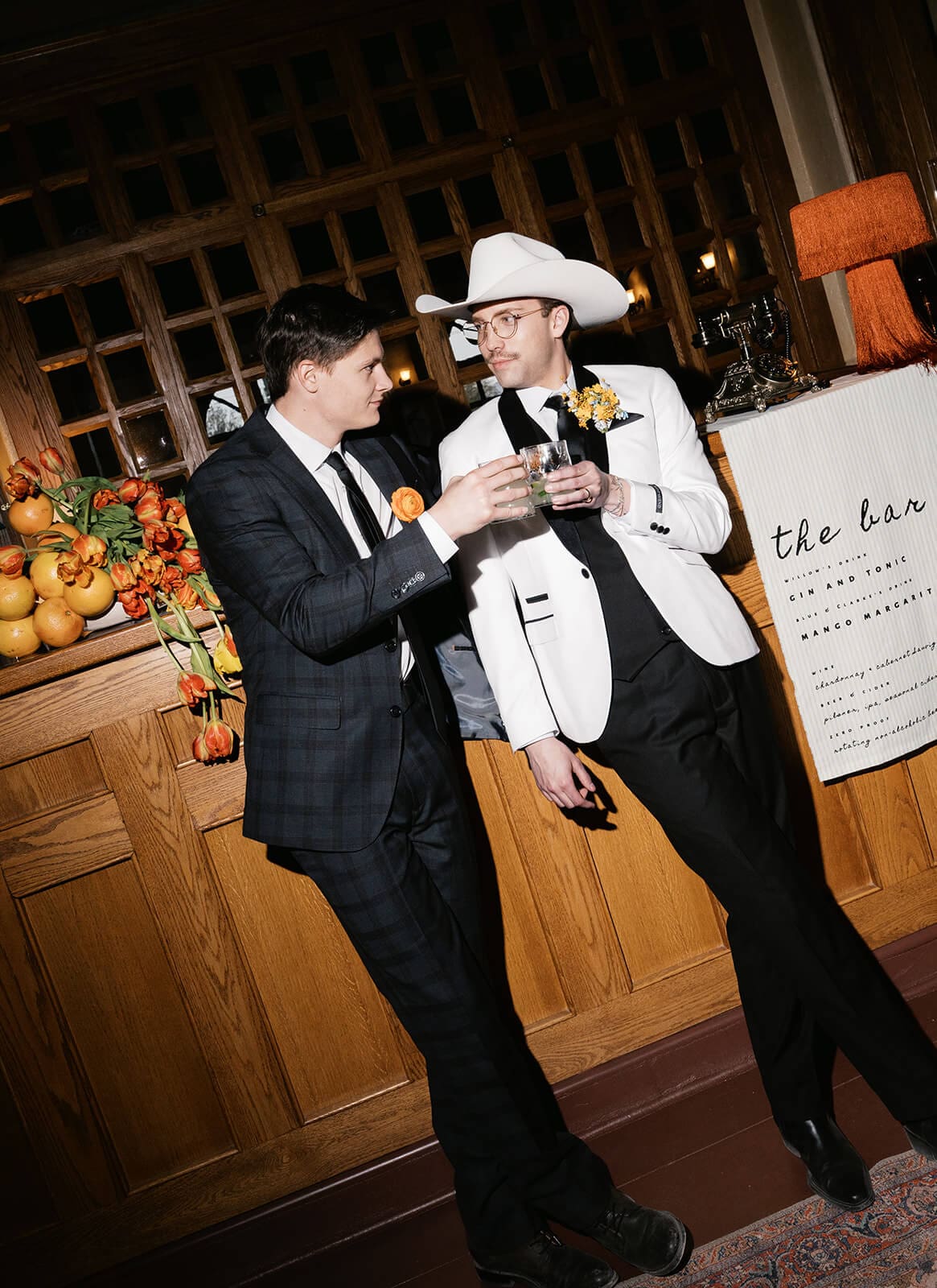 Two groomsmen toasting their glasses together near the same citrus-decorated bar, a hand-lettered bar menu visible behind them
