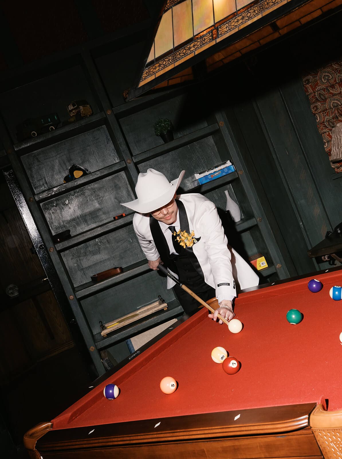 Groomsman in a white cowboy hat and white dinner jacket leaning over a red felt pool table lining up a shot, dark wood shelving in the background
