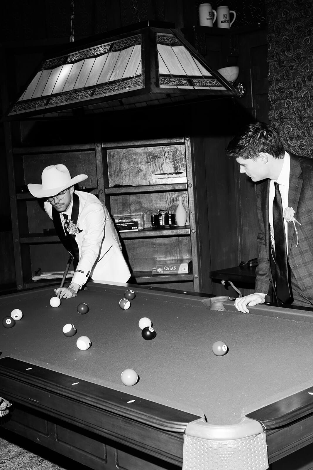 Black and white photo of two groomsmen in tuxedos playing pool under a stained glass swag lamp, one in a white cowboy hat leaning over the table lined with dark wood shelving
