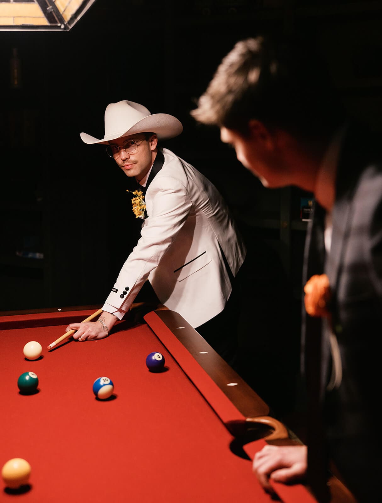 Groomsman in the white cowboy hat taking a shot at the red pool table while his groom looks on from behind