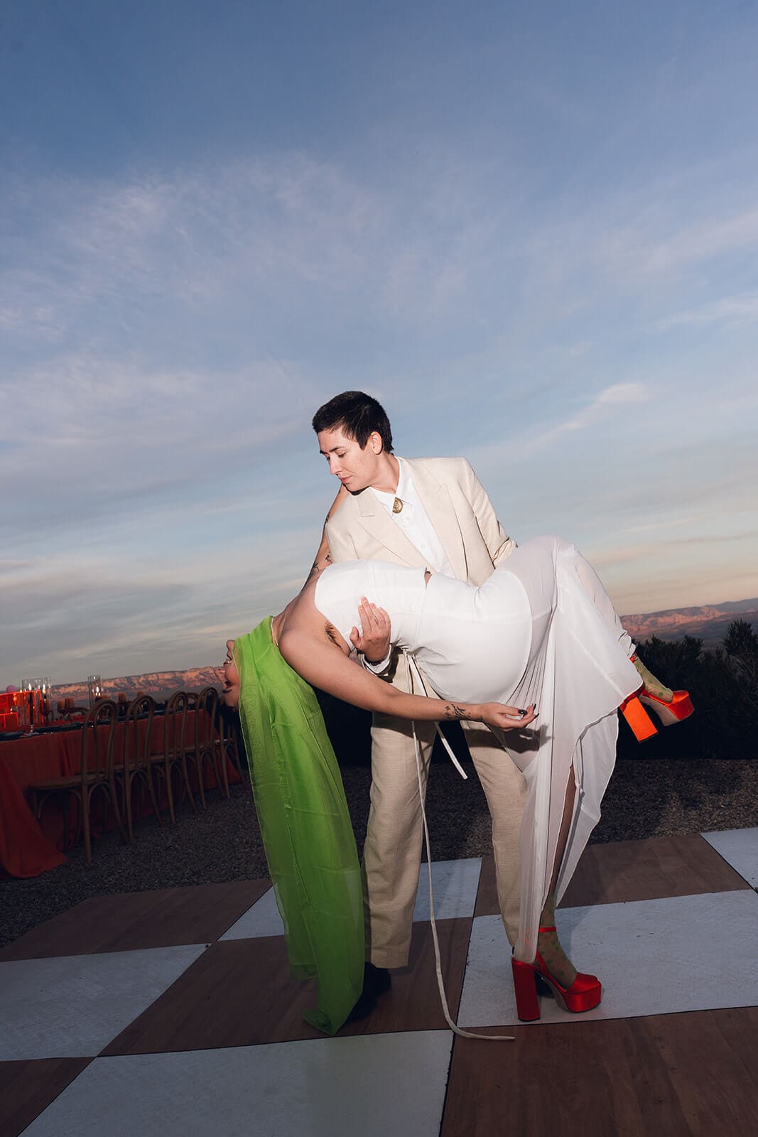 Person in a tan suit carrying the bride, who wears a white gown and red heels, across a black and white checkered dance floor under a soft pink and blue dusk sky
