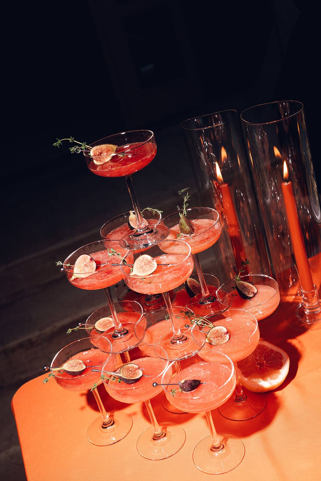 Tiered display of orange cocktails in coupe glasses garnished with citrus, arranged beside tall lit taper candles on an orange tablecloth
