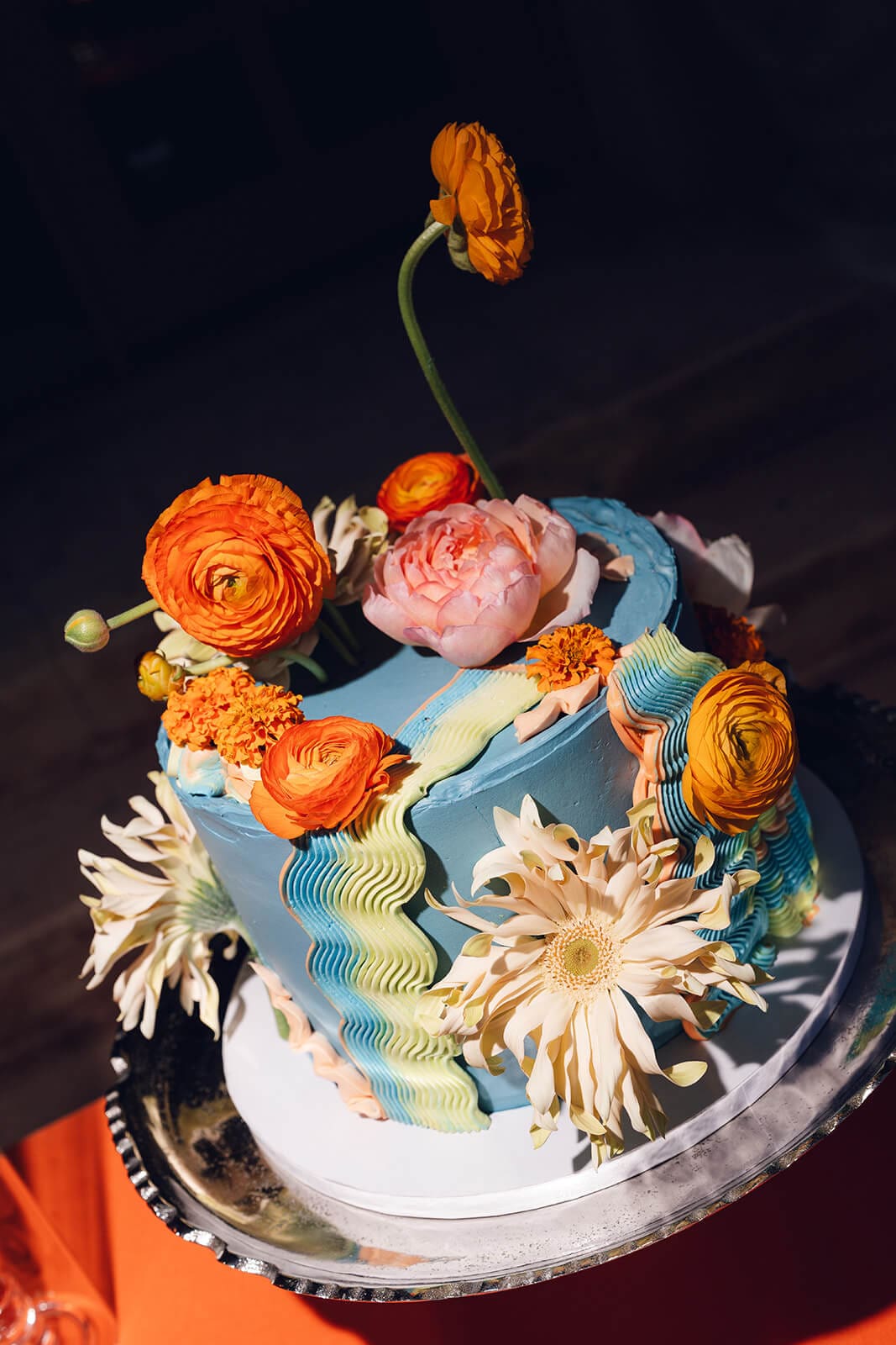 Two-tiered wedding cake with wavy blue buttercream detailing, topped with orange ranunculus, peach roses, and white daisies, one flower stem rising above the rest
