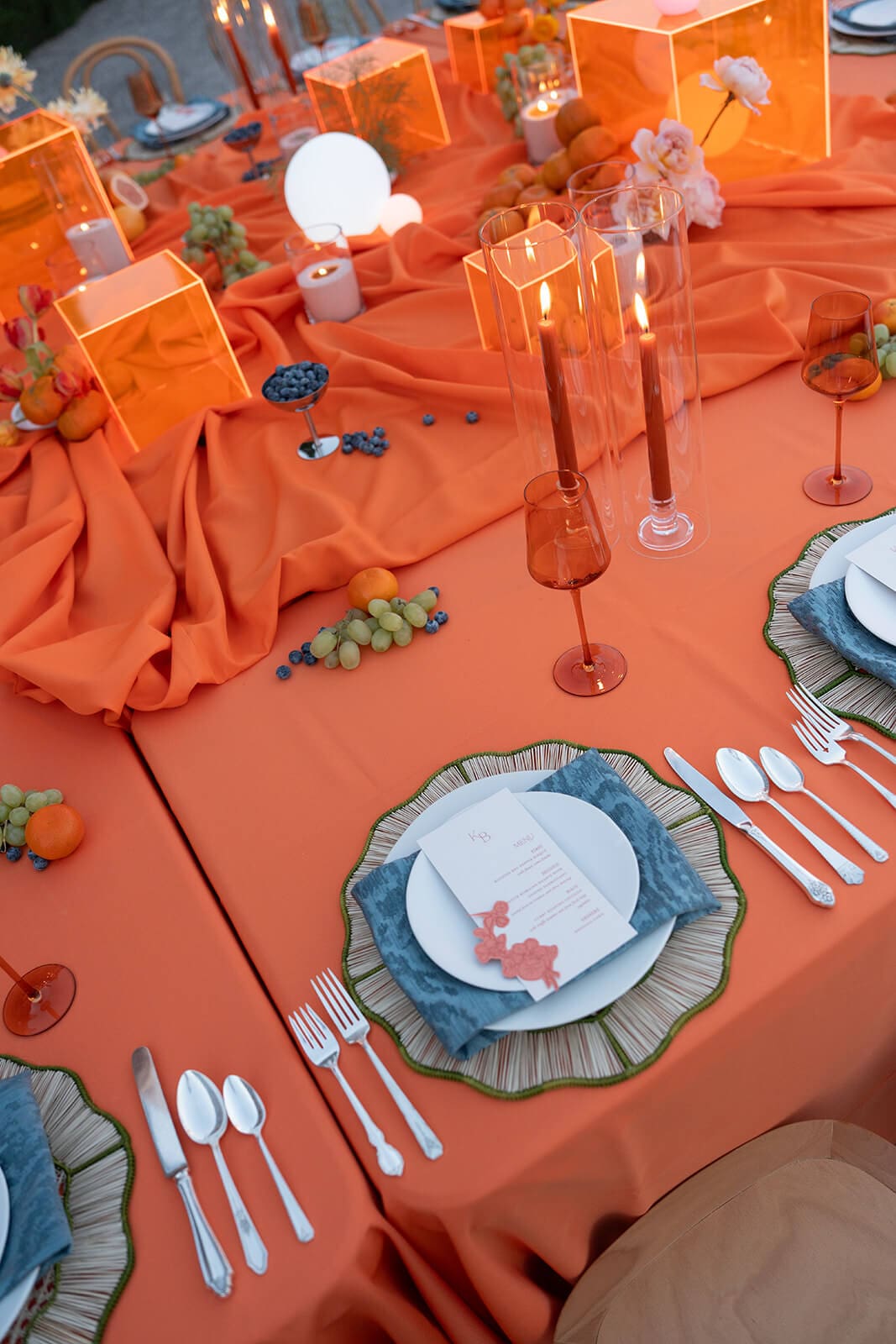 Close-up of an orange table setting with blue floral-rimmed plates, fresh citrus, small orange vessels, and lit taper candles at The Little Daisy wedding venue