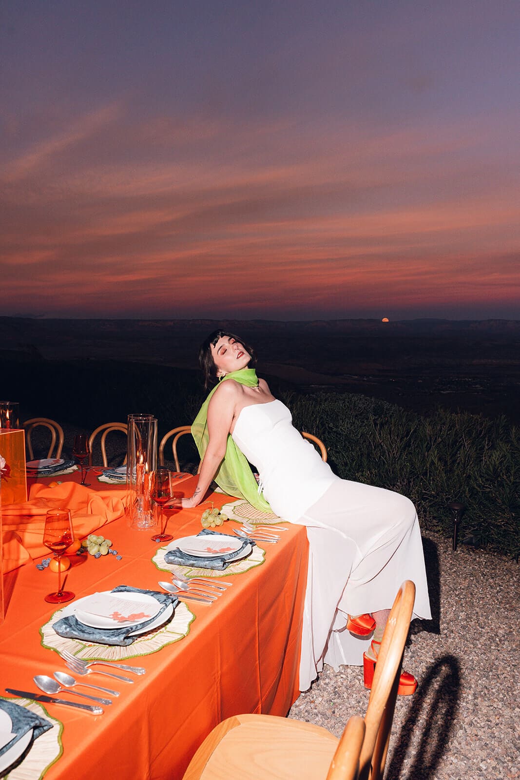 Bride sitting on the edge of a table at sunset, her green sash trailing, as a deep pink and orange sky stretches over the mountains behind her
