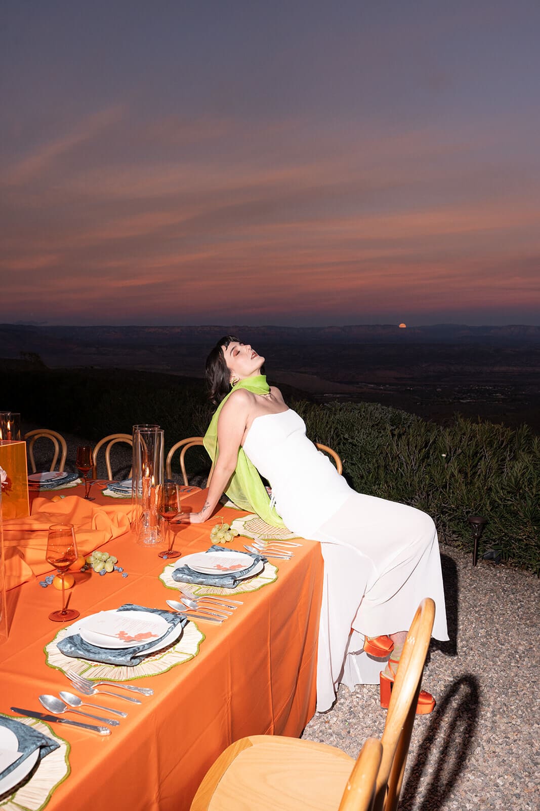 Bride sitting alone at an orange-draped table at dusk, mountains fading into the horizon behind her at The Little Daisy wedding venue.