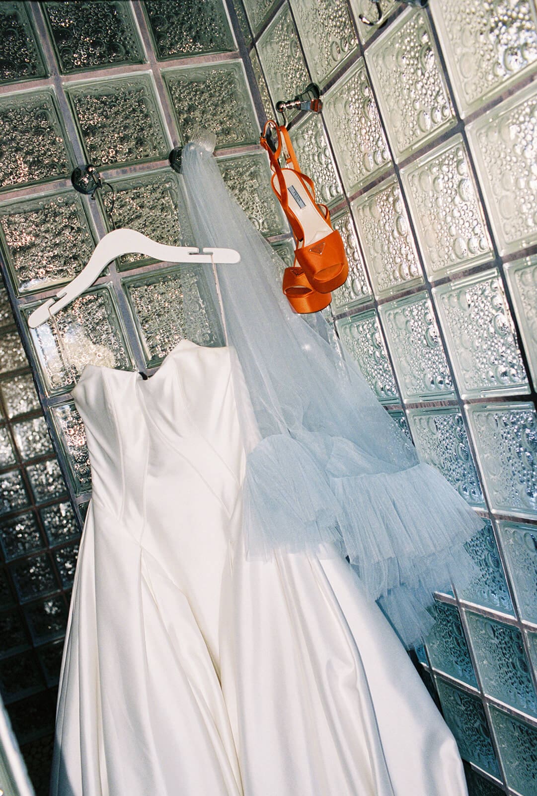 White satin wedding gown and pale blue tulle veil hanging against a glass block wall, with a pair of orange platform heels dangling from the hanger
