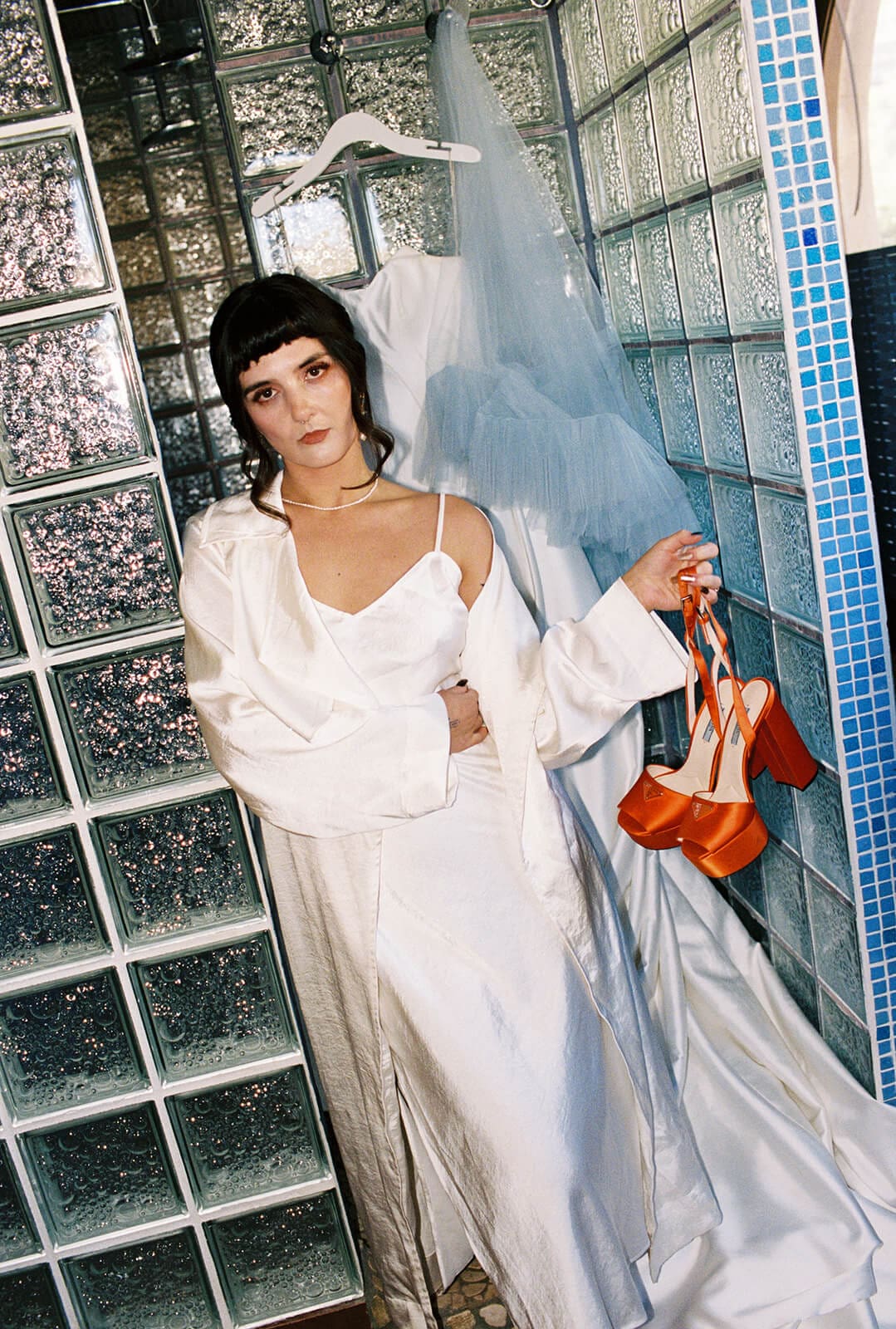 Bride in a white slip gown and flowing tulle veil posing against the glass block wall, one hand on her hip and the other holding her orange heels
