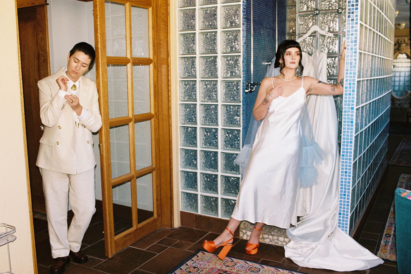 Bride in a spaghetti-strap white slip gown and tulle veil standing barefoot on tile in orange platform heels, glass block wall and Persian rug beneath her. Partner in a cream double-breasted suit adjusting his cufflink in a wood-framed doorway, eyes cast down
