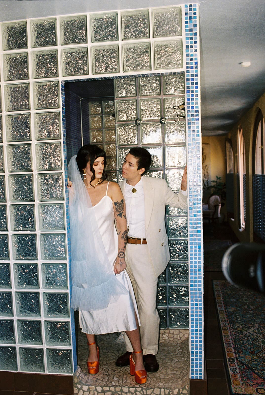 Bride and partner standing together in a mirrored glass block doorway, both dressed in white and cream
