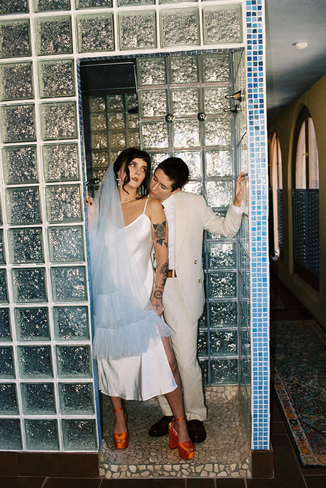 Couple embracing in the same glass block doorway, her orange heels peeking out from beneath her gown
