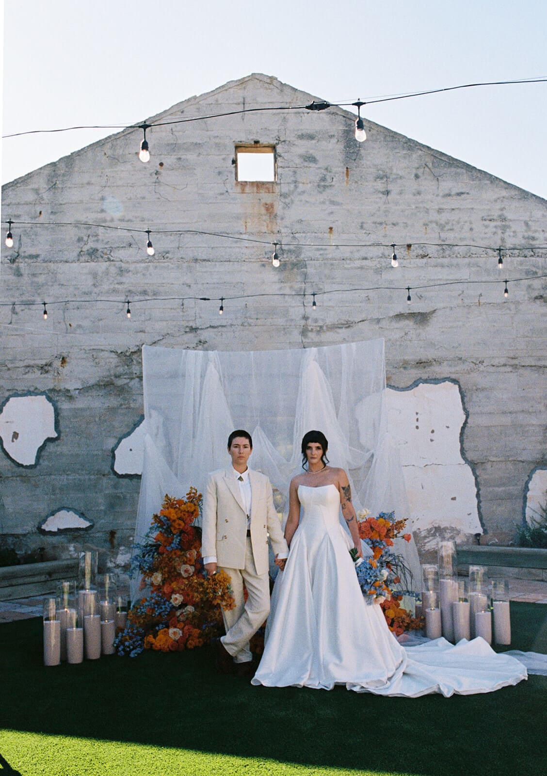 A couple stands against a weathered concrete wall with string lights overhead and an orange floral arrangement with pillar candles at their feet
