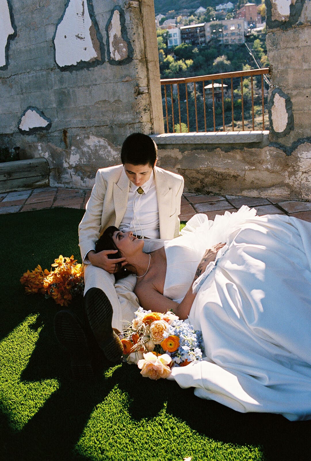 Couple sits on a green lawn surrounded by weathered stone architecture. Bride laying her head in her partner's lap, looking up at them, smiling.