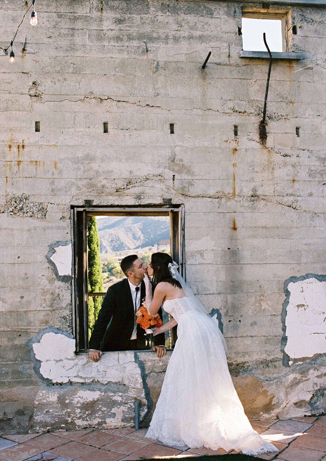 Bride and groom kissing through a rectangular stone window opening in a weathered wall, mountains and greenery visible beyond
