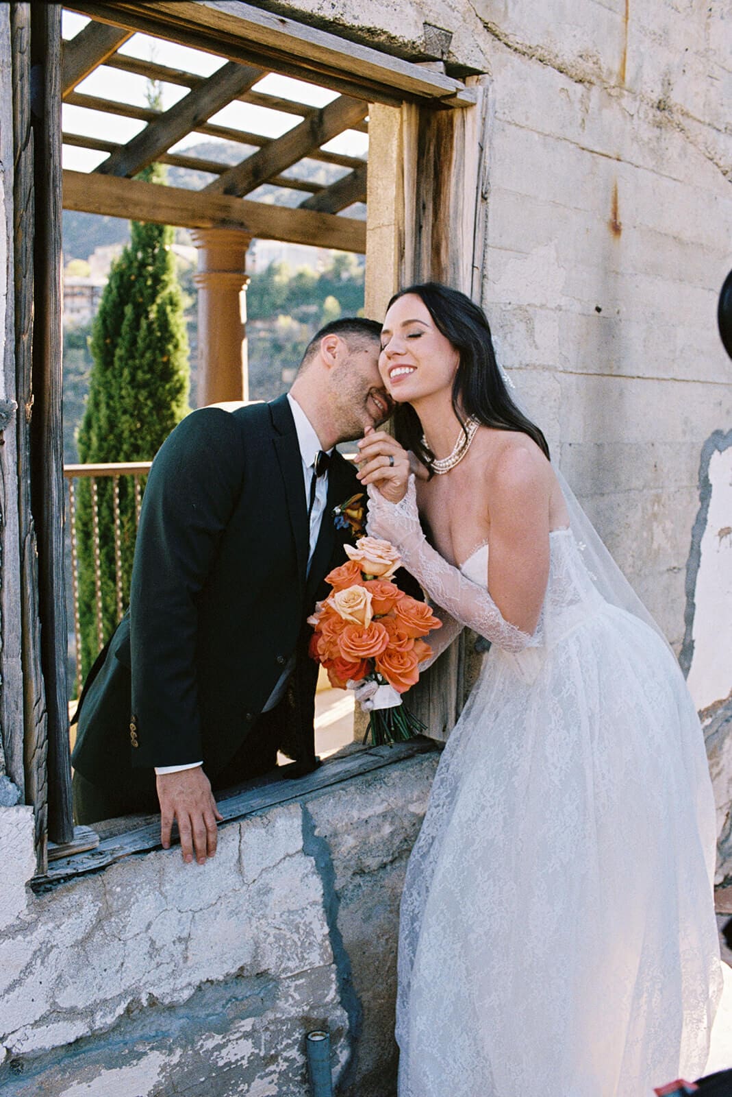 Groom kissing the bride's hand through the same stone window as she laughs, holding a bouquet of orange roses
