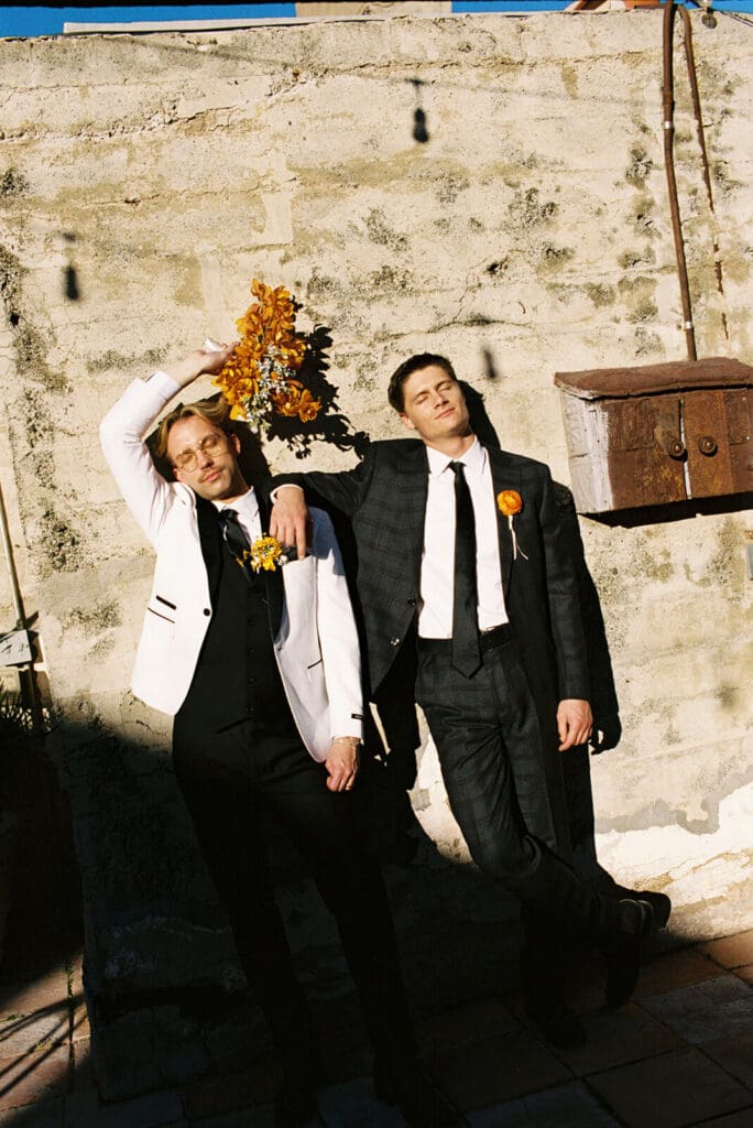 Couple in formal suits posing against a rustic stone wall, with one holding an orange floral arrangement overhead.