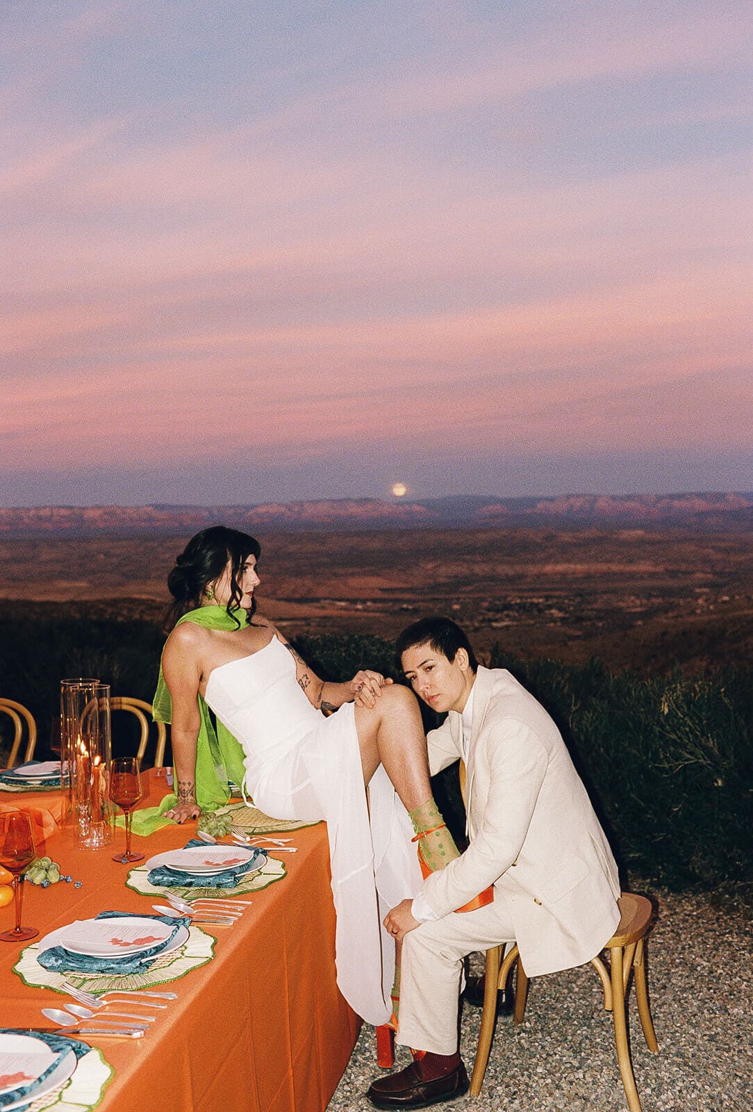 Couple seated at the edge of a long table, watching a full moon rise over the desert landscape at dusk during a reception dinner at The Little Daisy wedding venue
