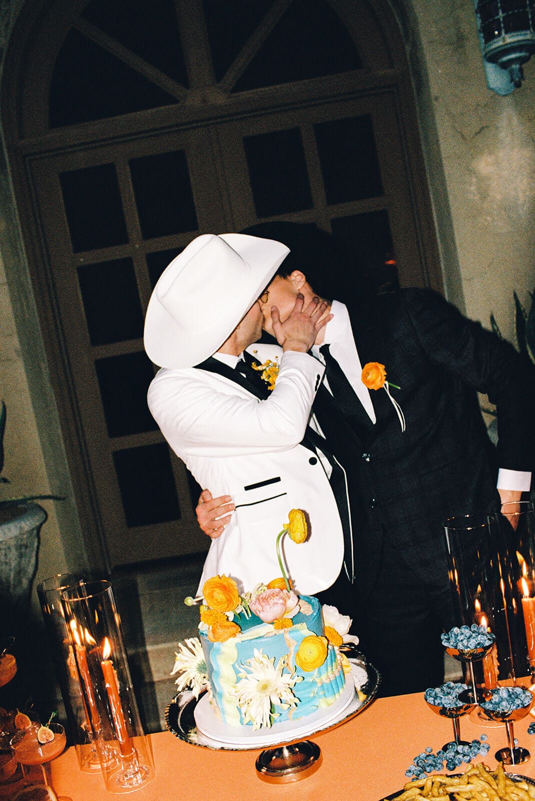 Two grooms kissing over their wedding cake at night, one in a white cowboy hat, surrounded by lit taper candles and orange florals
