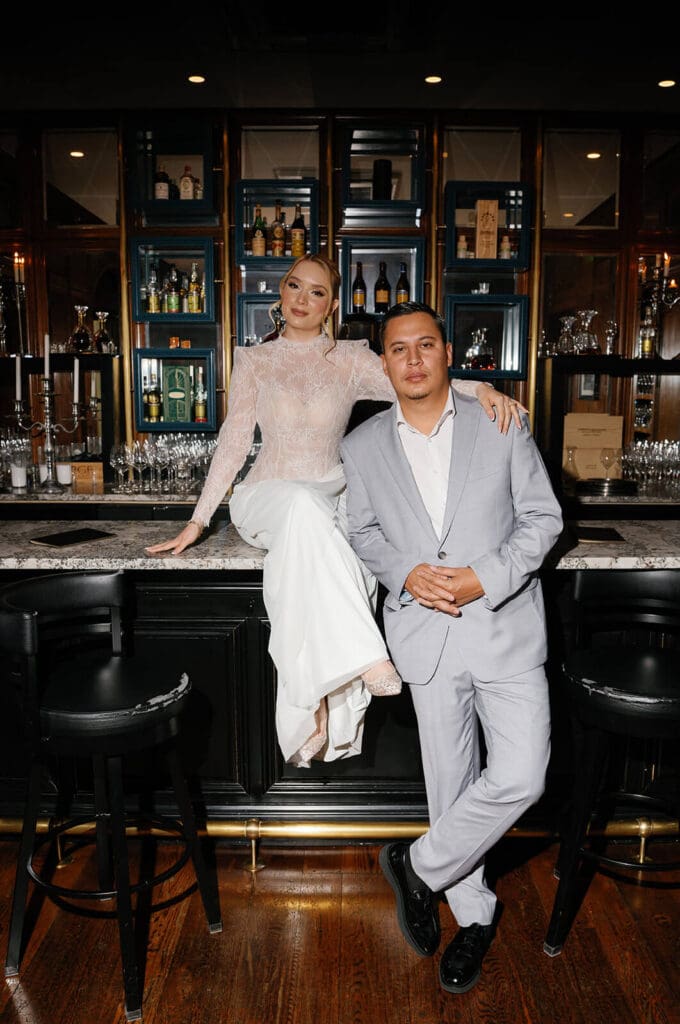 Newlyweds posing together at a dark wood bar, with the bride sitting on the countertop beside her partner.