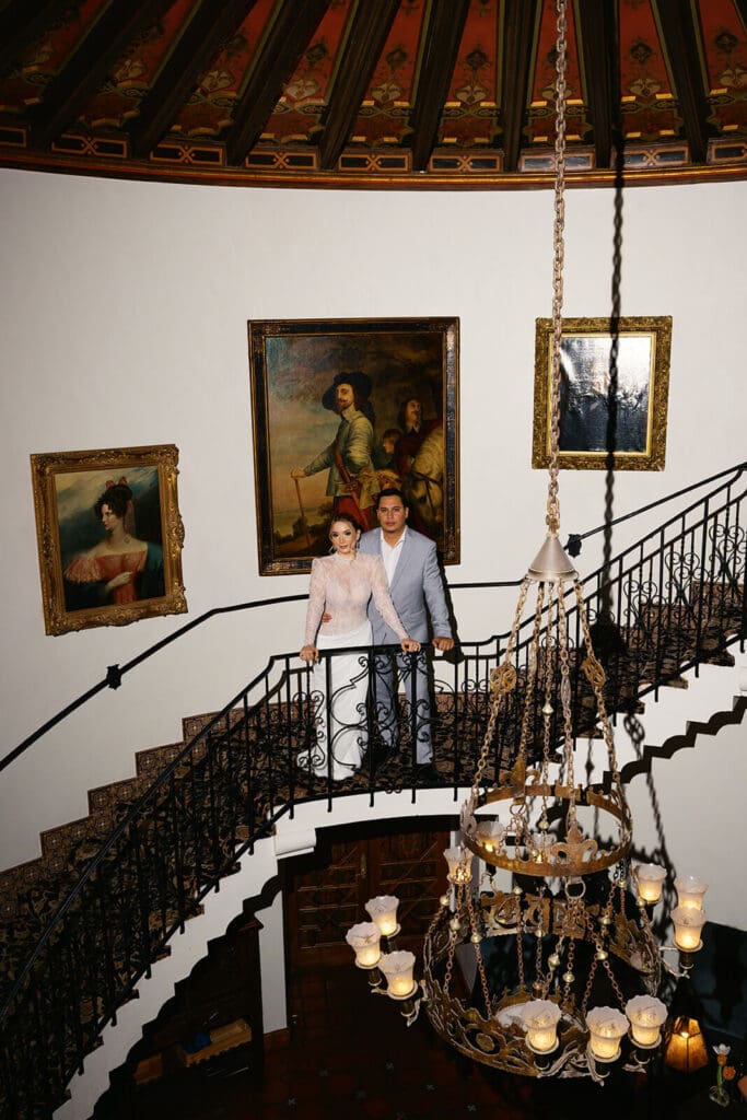 Couple standing on a grand staircase beneath a chandelier inside a historic venue filled with artwork.