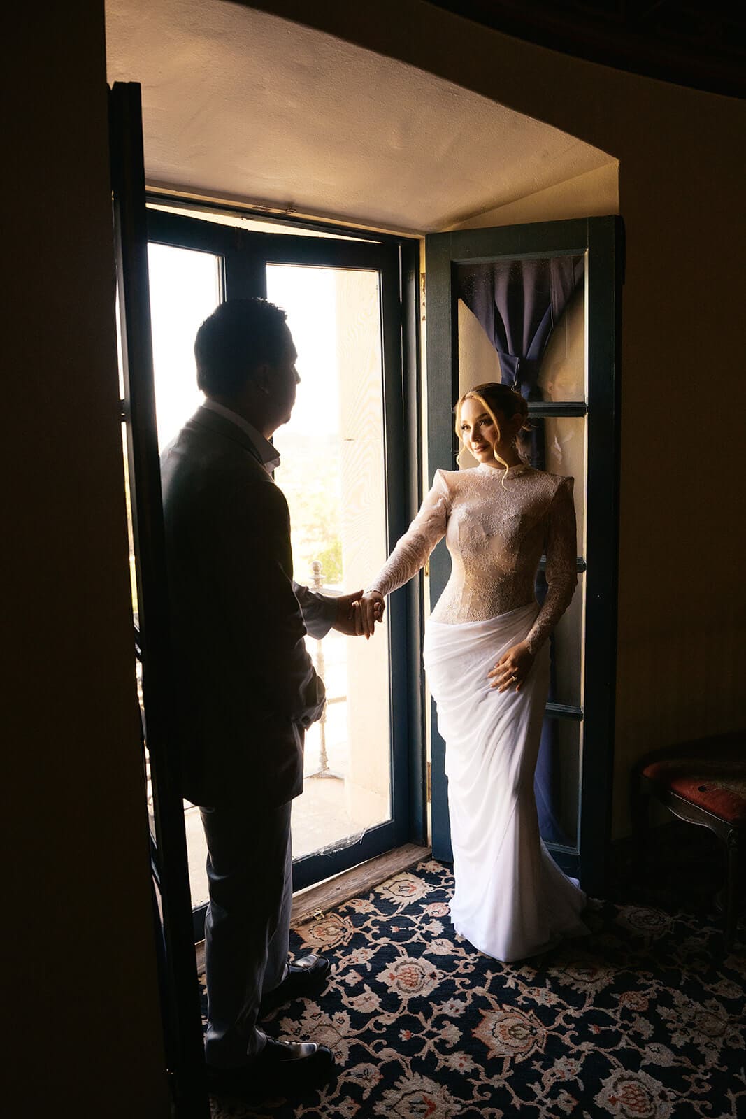 Couple holding hands while stepping through a sunlit doorway inside a historic mansion.