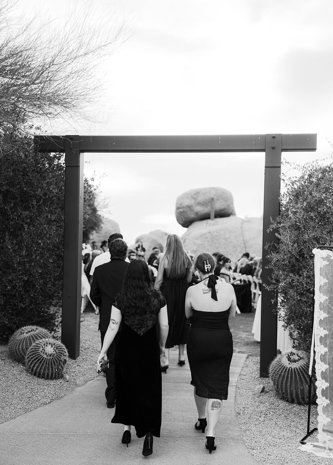 Black-and-white photo of wedding guests walking through a modern entryway toward an outdoor desert ceremony.