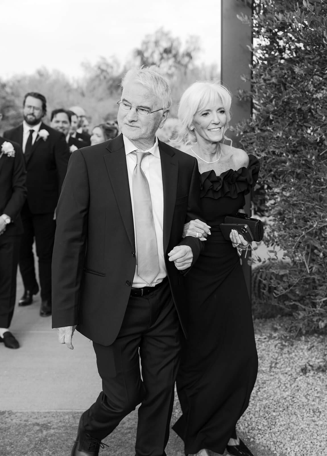 Black-and-white photo of an older couple walking arm in arm toward the wedding ceremony.