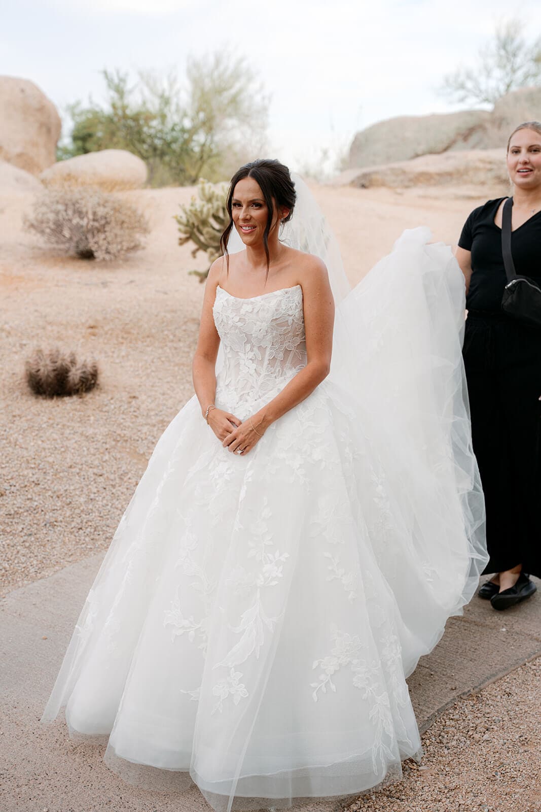 Bride in a lace wedding gown standing beside desert boulders while an attendant adjusts her train.