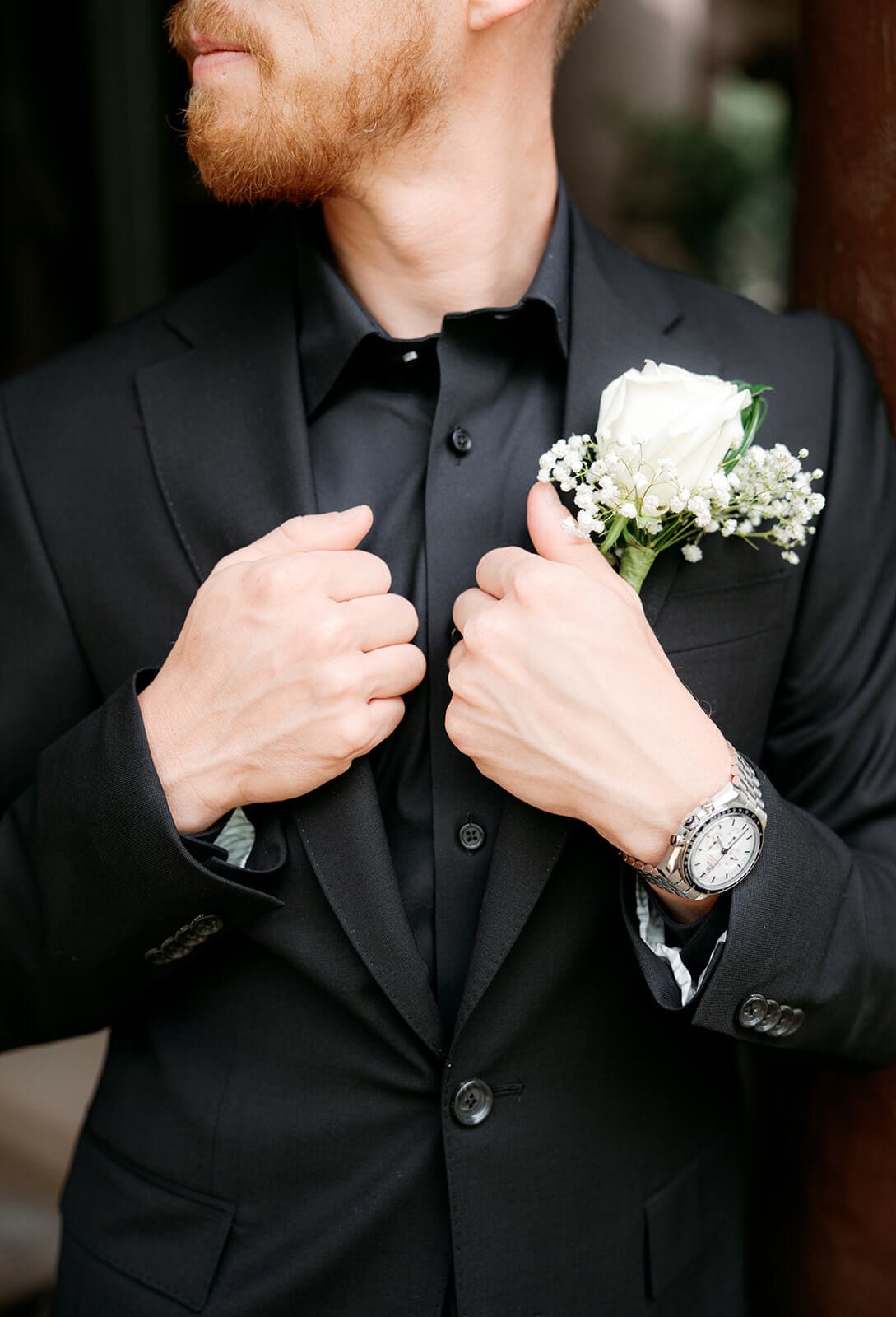 Close-up of the groom adjusting his black suit jacket beside a white boutonniere and wristwatch.